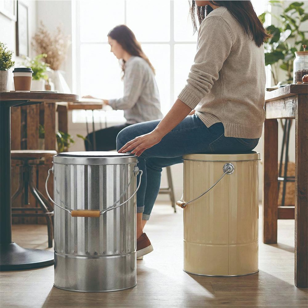 兩名女性坐在儲物桶上，一個是銀色帶木把手，另一個是米白色，室內有窗戶和桌子。