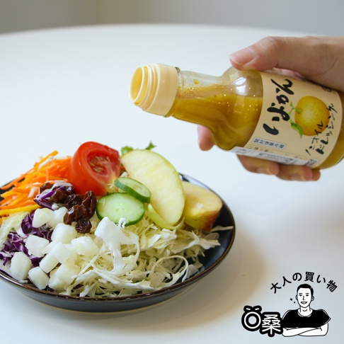 玻璃瓶裝無油柑橘風味沙拉醬，黃色醬汁從瓶口淋在色彩豐富的沙拉上，沙拉包含切片番茄、小黃瓜、蘋果、紫甘藍、紅蘿蔔絲和白色塊狀物。