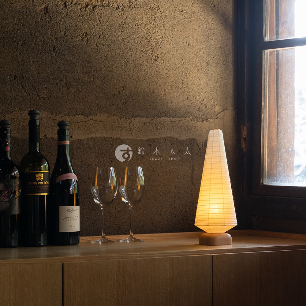 一個錐形紙質提燈，燈罩為米白色，底部為淺木色底座，發出暖黃色光芒，擺放在木質桌面上，旁邊有兩瓶紅酒和兩個酒杯。