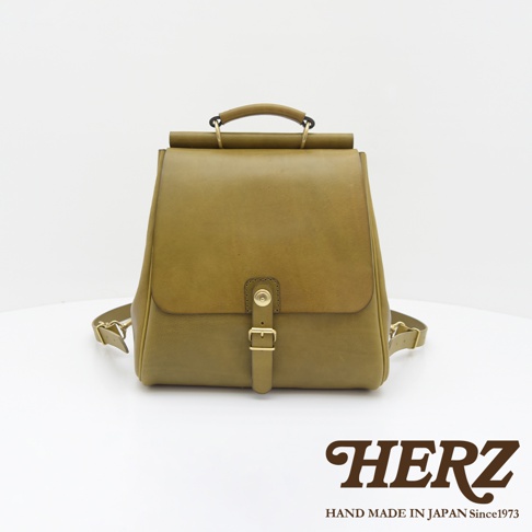 一款橄欖綠的皮革後背包，具有屋瓦造型的翻蓋設計，配有金色金屬扣環和皮革揹帶，展現職人手作質感。