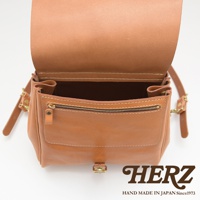 焦糖色皮革後背包，帶有拉鍊前袋和金屬釦，內部空間可見，展現職人手作質感。