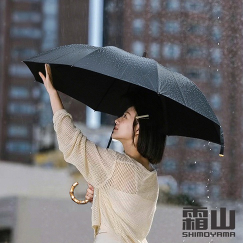 一位短髮女性在雨中撐著一把黑色摺疊傘，這把傘配有獨特的復古風格竹製彎柄，畫面右下方印有霜山SHIMOYAMA品牌字樣，呈現出優雅的雨天氛圍。