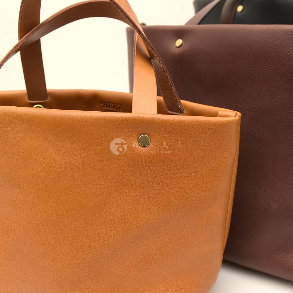 一只棕褐色的職人手作皮革極簡托特包，配有同色系皮革提把，包身前方有一個金屬卯釘。背景是一只深棕色的同款托特包。