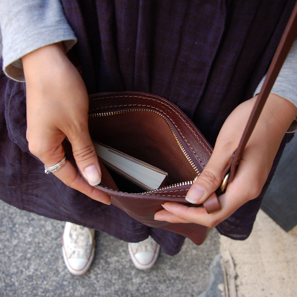 職人手作皮革Organ肩背小方包，深棕色光滑皮革，內有淺色小書，搭配銀色戒指，由女性雙手拿取。