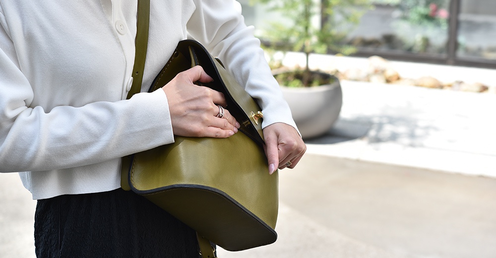 一位女士手持橄欖綠皮革翻蓋迷你後背包，露出背包的頂部翻蓋和肩帶。