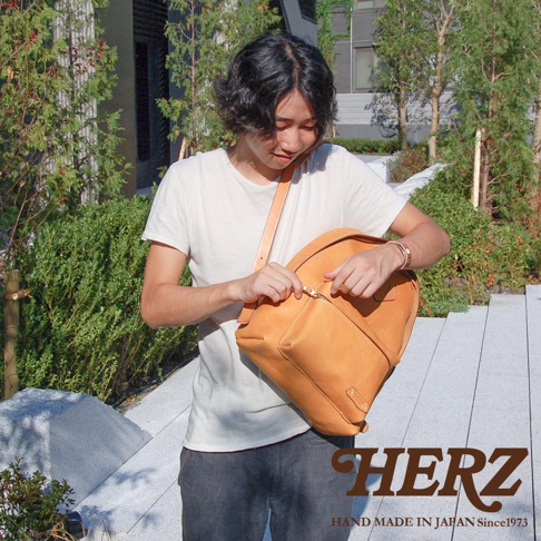 職人手作皮革休閒風後背包 R-93，展示於戶外環境。背包為亮棕色皮革材質，設計簡約，設有拉鍊開口及前袋，由一位男性揹負，正在拉動背包拉鍊。