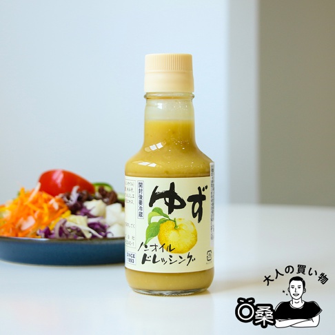 玻璃瓶裝無油柚子風味沙拉醬，醬汁呈淡黃色。瓶身標籤為淺黃色，印有日文「ゆず」和柚子圖案。背景是藍色盤子裡的沙拉，包含切絲的胡蘿蔔、紫甘藍和番茄。