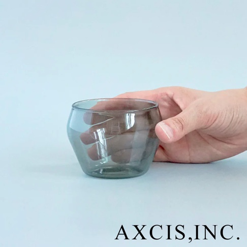 一手握著一個煙灰色、透明、帶有復古圓潤造型的玻璃杯，杯身有波浪紋理。