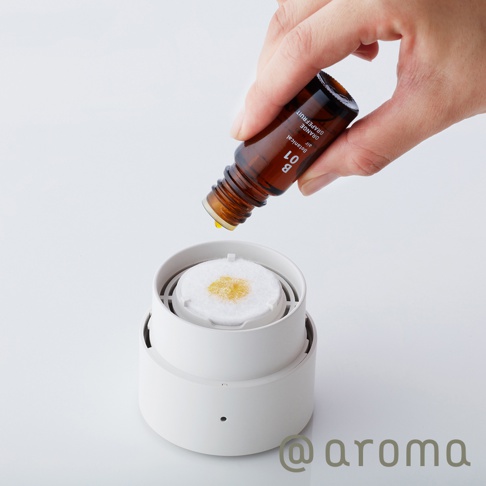 手持一瓶琥珀色玻璃瓶裝的精油，將黃色精油滴入白色圓形香氛機的白色吸附棉墊上。