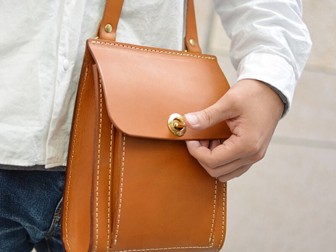焦糖色厚實皮革單肩包，帶有金色旋轉鎖扣和白色縫線細節，由手持展示。