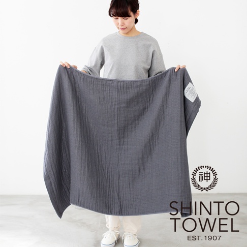 一位女士展示著一條深灰色、2.5層紗材質的浴巾，浴巾呈現自然的皺褶紋理，左上角有一個白色標籤，右下角有「SHINTO TOWEL EST. 1907」字樣及品牌標誌。