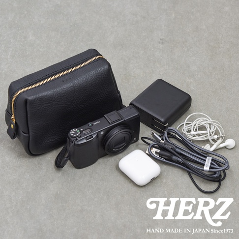 黑色皮革手作收納包放置於灰色地面，周圍陳列相機、白色耳機盒、充電器及數據線等隨身物品，底部印有日本職人品牌HERZ商標與成立年份。