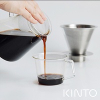 透明玻璃咖啡壺傾倒咖啡液至玻璃咖啡杯，背景為銀色不鏽鋼濾網濾杯。