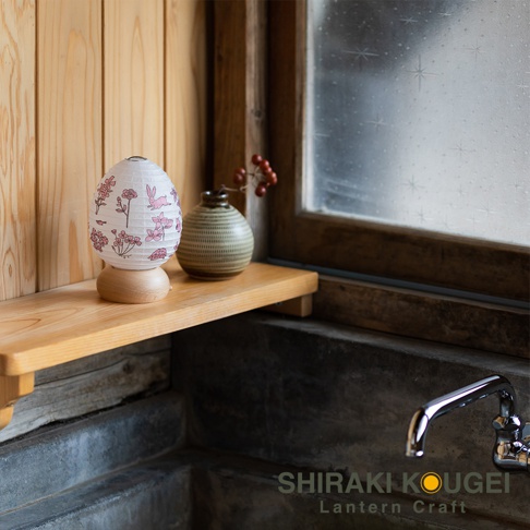 一個裝飾提燈，燈罩為白色和紙材質，印有粉色花卉和兔子圖案，底部為木質底座。提燈旁有一個綠色條紋紋理的陶瓷花瓶，瓶中插有帶有紅色漿果的枝條。