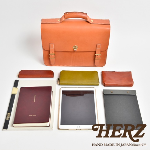 橘色皮革公事包，附有兩條搭扣和金色鎖扣，側邊擺放著棕色筆袋、橄欖綠長夾、橘色小化妝包、紅色筆記本、平板電腦及灰色記事板。