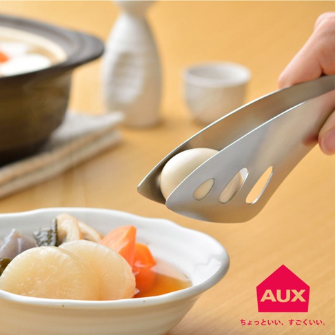 銀色不鏽鋼料理夾夾著一顆水煮蛋，夾身有三個長條形孔洞，背景是裝有燉菜的白色碗以及日式酒器。