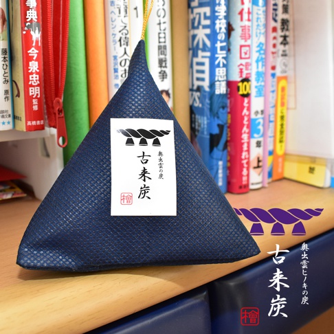一個深藍色織紋布料製成的三角形香氛袋，放置在書架前。袋子中央貼著白色標籤，印有黑色書法字體古來炭以及代表神聖的注連繩圖案和紅色印章。畫面右下方重疊顯示了相同的注連繩圖案、古來炭書法字及紅色印章標示，整體呈現日式傳統工藝的簡約質感。