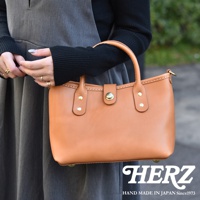 職人手作皮革迷你托特包，採用光滑棕色皮革，配有金色金屬扣和鉚釘。包包有兩個短提把，並透過車縫線裝飾，整體呈現簡約實用的風格。