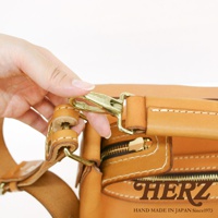 一只手正操作著一個黃銅色的鉤扣，連接到一個 tan 色皮革製成的波士頓包。包包有明顯的縫線細節和一個拉鍊。