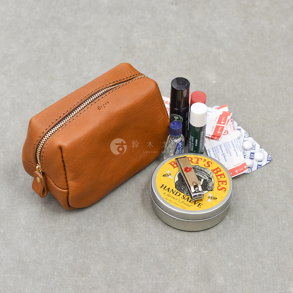 一個焦糖棕色的日本HERZ品牌Organ系列皮革拉鍊收納包，放置在灰色平面上，包旁散落著生活小物，包括一罐伯特小蜜蜂護手霜、指甲剪、幾管護唇膏與眼藥水，以及幾片包裝藥錠和OK繃。