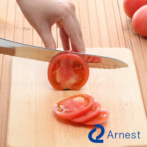 手持不鏽鋼麵包刀，正在切一片紅色的番茄，刀刃呈鋸齒狀。番茄被切成數片，放置在木質砧板上，背景是木紋桌面。