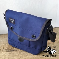 藍色純棉帆布雙扣郵差包，帶有灰色滾邊、黑色皮革裝飾和可調節黑色肩帶。包蓋上印有品牌標誌。