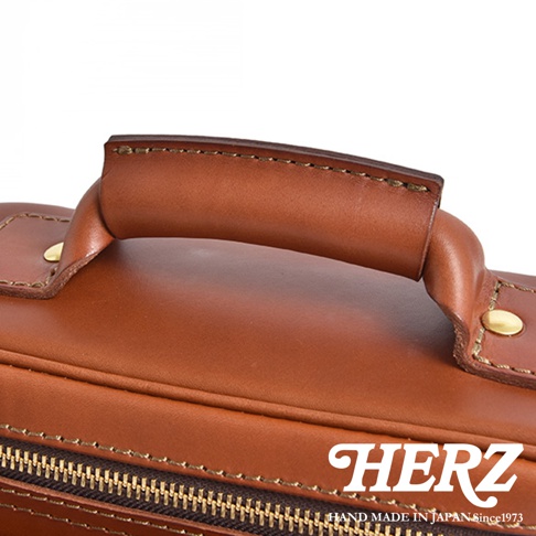 HERZ 職人手作皮革厚實相機包，特寫皮質提把和拉鍊細節，右下角有 HERZ 品牌標誌與「HAND MADE IN JAPAN Since1973」文字。