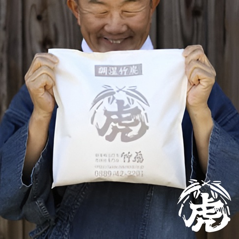 一位男子手持白色編織袋，袋上印有灰色「虎」字圖案及日文文字「調濕孟宗竹炭」。