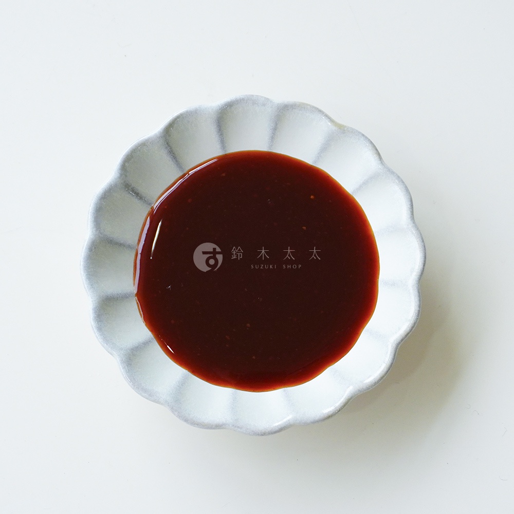白色花瓣造型的陶製小碟子置於純白背景中央，碟中盛裝著深褐色的朝紫大蒜味噌萬用調味醬，醬料質地濃稠且帶有細微顆粒，盤面中心印有鈴木太太的品牌標誌。