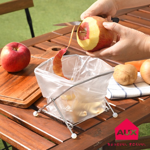 銀色金屬支架支撐著透明塑膠袋，袋內裝有蘋果皮和馬鈴薯。一名女子正用刀削蘋果，蘋果皮剝落進垃圾袋。桌面上還有一個紅蘋果、馬鈴薯、洋蔥和紅蘿蔔。