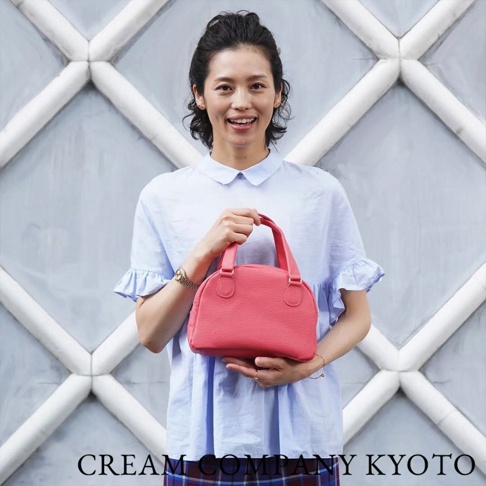 一位女子手持一個珊瑚紅色、荔枝紋皮革的迷你手提包，包身呈半月形，配有雙提把。