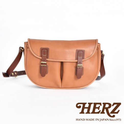 棕褐色皮革製的傳統釣魚包，翻蓋設計，配有雙皮帶和黃銅扣環。包身線條圓潤，縫線精緻，肩帶可調節。