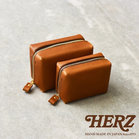 兩個職人手作皮革箱型收納包，一前一後擺放，皆為棕褐色，配有金色拉鍊和皮革拉環。