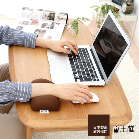棕色布面減壓護腕枕，放置在木質桌面上，支援使用滑鼠的手腕，一旁有筆記型電腦和雜誌。