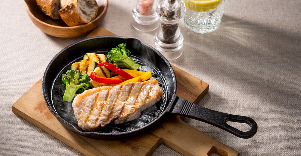 黑色的鑄鐵波浪平底鍋中盛有煎烤的豬排、綠色花椰菜、櫛瓜片、紅黃甜椒，鍋底有烤痕，散發著微微的熱氣。