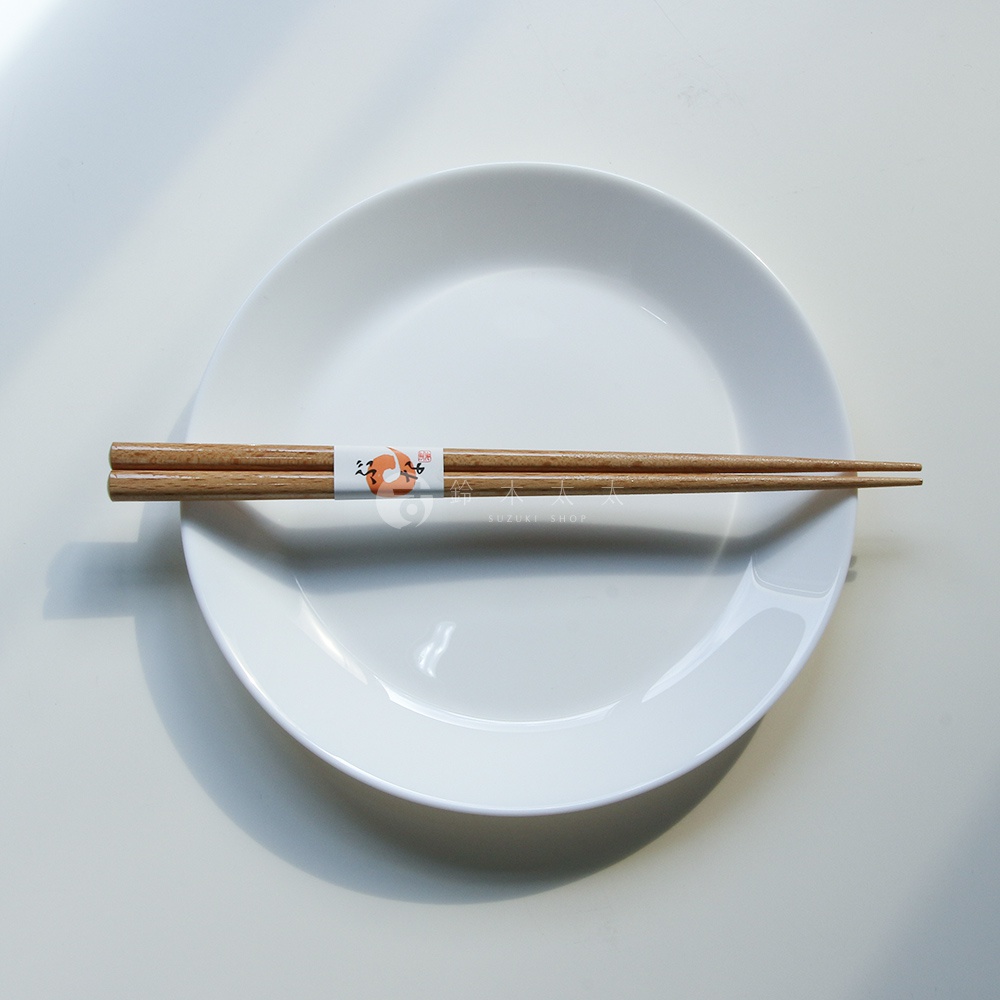一雙天然原色木筷，表面有細緻的紋理，筷頭尖細，中段有白色標籤，標籤上印有日文文字及圖案，筷子置於白色圓盤上。