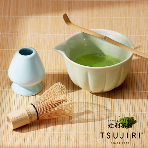 竹製抹茶刷、淡綠色片口碗及竹匙擺放在竹簾上，旁邊有一座淺藍色茶筅立，碗中裝有鮮綠色的抹茶，右下角印有辻利茶舖品牌標誌及相關文字。