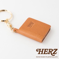 職人手作皮革書本造型鑰匙圈，淺棕色皮革，附有金色金屬鑰匙圈和扣環，皮革表面壓印有「HERZ MADE IN JAPAN」字樣。