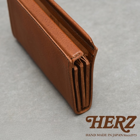 由日本品牌HERZ手工製作的棕色皮革長夾，從側面特寫展示其多層次的風琴式結構與細膩的縫線細節，下方標示有HERZ品牌名稱及手作工藝資訊。