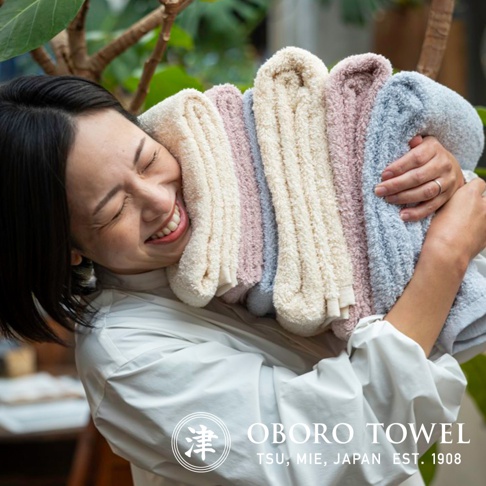 一位女子抱著一疊柔軟的有機棉浴巾，浴巾顏色包含米白色、淡粉色和淺藍色，質地蓬鬆，呈現出舒適的觸感。