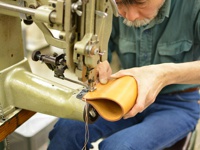 一位工匠正使用工業縫紉機縫製一個淺棕色的皮革筆筒。皮革製品的邊緣有縫線，顯示出精緻的工藝。