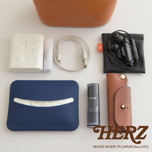 深藍色皮革收納包內含面紙，旁邊有白色行動電源、護唇膏、充電線、黑色耳機與皮製鑰匙圈。