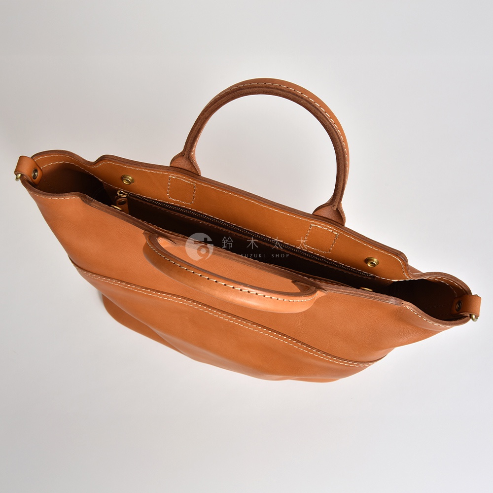 職人手作皮革大容量兩用托特包，呈現溫潤的焦糖棕色，以堅韌的皮革材質和精緻的白色縫線製作，包身線條簡潔，配有圓潤的提把，展現經典風格。