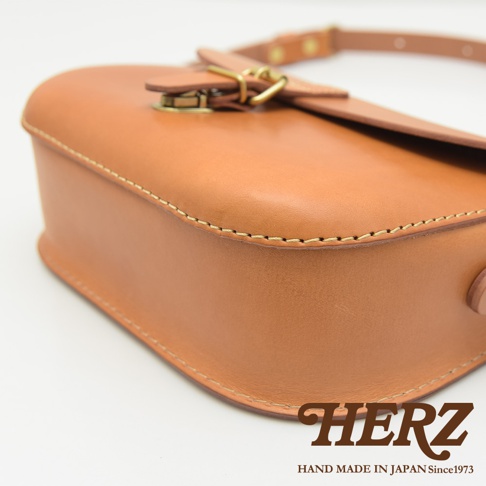 職人手作皮革經典馬鞍包，淺棕色皮革，包身縫線細緻，搭配金色金屬釦與皮質揹帶。