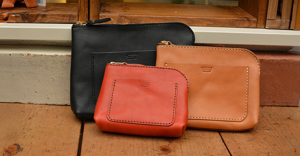 三款職人手作皮革小物收納包，分別為黑色、橘紅色及棕褐色。包款材質為皮革，皆有拉鍊開合，並附有外層口袋，縫線明顯，質感佳。