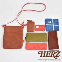 職人手作皮革直式隨身單肩包，棕色皮革主體，搭配紅色細節。一旁展示其他皮革配件，包含棕色鑰匙包、紅色巴士造型卡夾、橄欖綠長夾、珊瑚粉筆袋，以及一本棕色皮革封面筆記本，和藍色印花手帕。