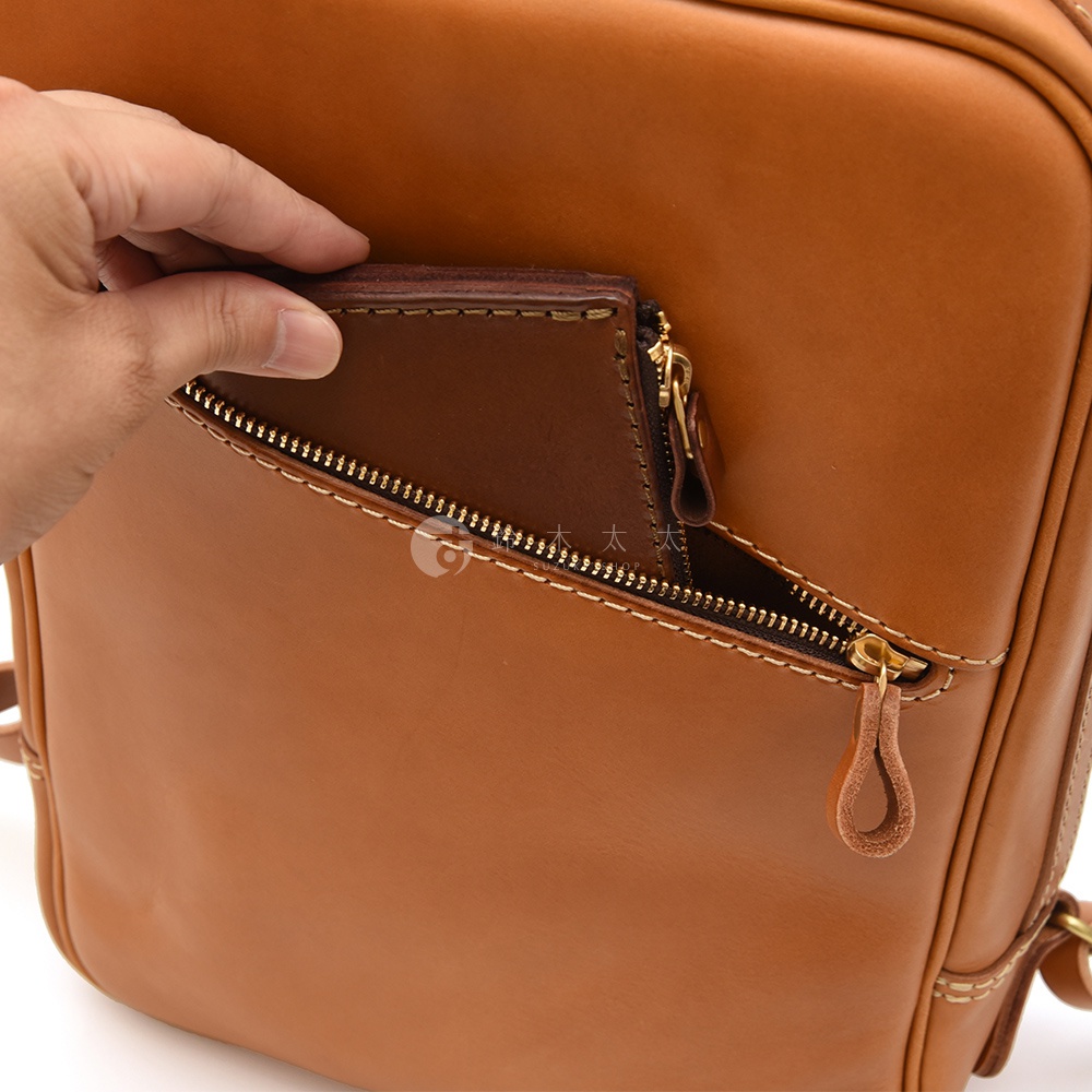 職人手作皮革長方型多口袋後背包 R-17，展示棕色皮革材質，一個手將深棕色錢包放入後背包前方拉鍊口袋，拉鍊為金色，縫線清晰。