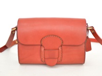 一款橘紅色皮革製成的寬扣帶馬鞍包，包身有明顯的縫線裝飾，正面有一個突出的皮革搭扣，側邊懸掛著一個皮革吊牌。