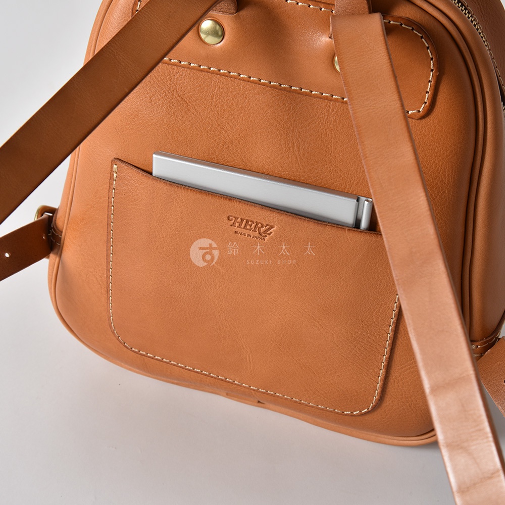 駝色皮革迷你後背包，正面設有立體口袋，內有薄型金屬物品，背包肩帶為皮革材質，車縫線清晰。