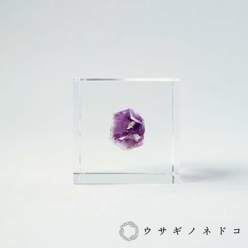 透明立方體中懸空放置著一塊紫水晶原礦，呈現不規則的塊狀，表面有紫色晶體和部分白色雜質。